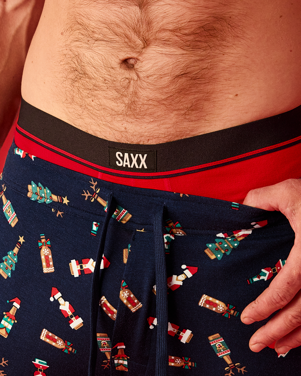 Close-up of a man wearing SAXX men's boxer briefs. The underwear features a dark navy blue base with a festive Christmas print of Santa Claus, elves, and snowman figures, a solid red waistband, and a black center band with the white SAXX logo.