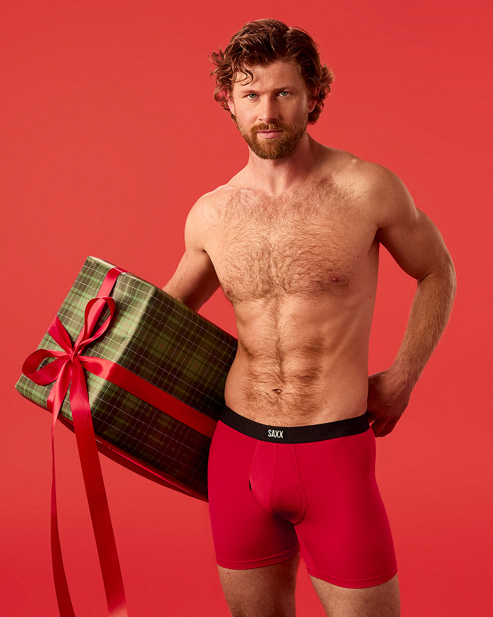 A shirtless man with a beard wearing red boxer briefs stands against a solid red studio background, holding a large wrapped gift box with a red ribbon. The waistband of the underwear shows the brand name 'SAXX'.