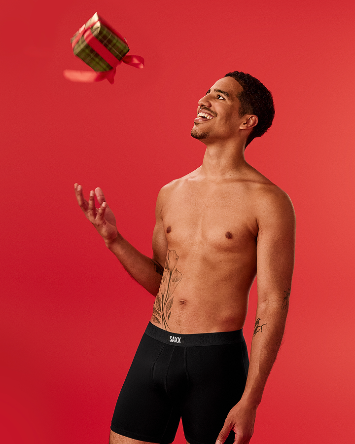 A shirtless man wearing black boxer briefs with a visible SKYN brand waistband, standing against a solid red studio background and reaching toward a small wrapped gift box floating in the air.