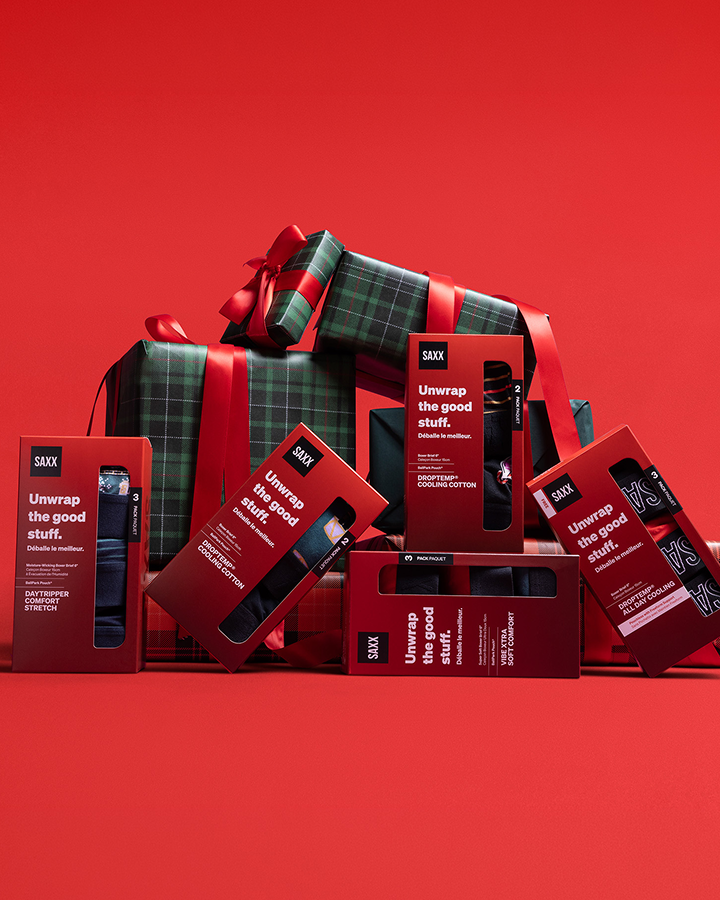 A styled arrangement of SXXX gift boxes and wrapped presents on a solid red background. The packaging is deep red with white text reading 'Unwrap the good stuff.