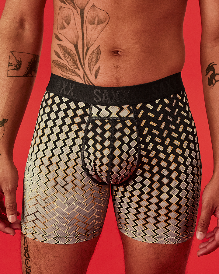 A close-up of a male model wearing SAXX boxer briefs with a black and beige geometric zigzag pattern, set against a solid red background.