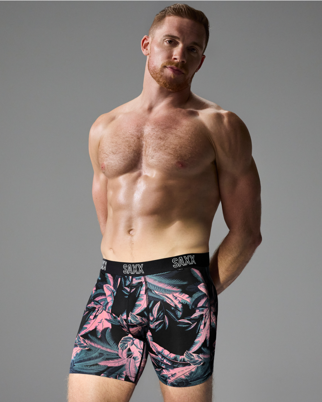 A shirtless male model wearing boxer briefs with a black tropical leaf pattern in pink and teal, posed against a solid gray studio background. The brand name 'SAXX' is visible on the waistband.