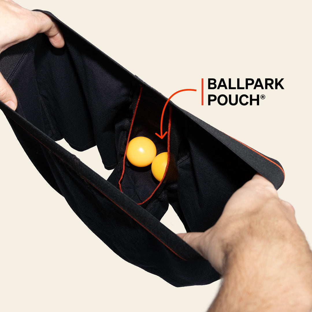 Black boxer briefs opened to show the interior Ballpark Pouch holding two yellow balls, with an arrow labelling the BallPark Pouch