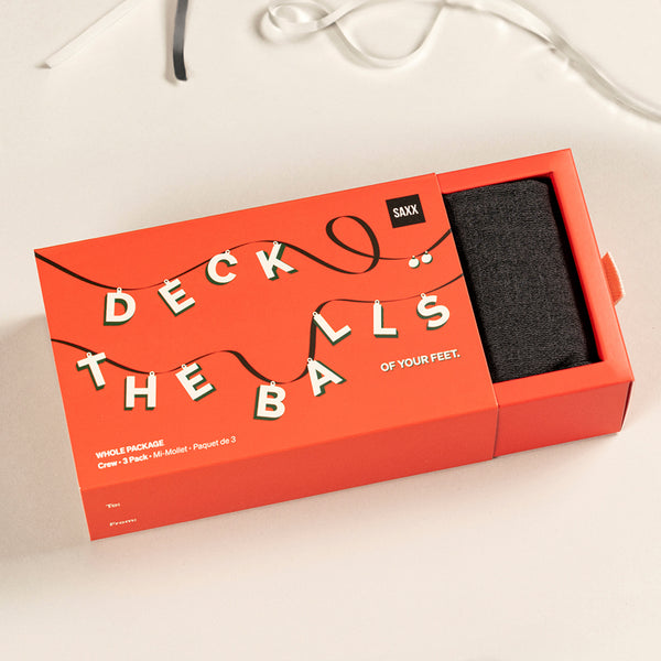 Red box with phrase "Deck the Balls" on white table being opened to reveal black socks