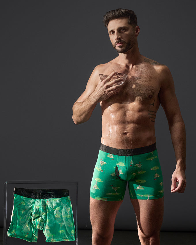 A shirtless man modeling green patterned boxer briefs with a black waistband, standing against a dark gray studio background. A smaller inset at lower left shows the boxer briefs laid flat.