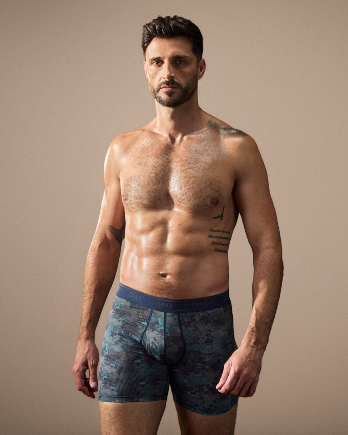 A shirtless man modeling blue camouflage boxer briefs against a plain beige studio background.