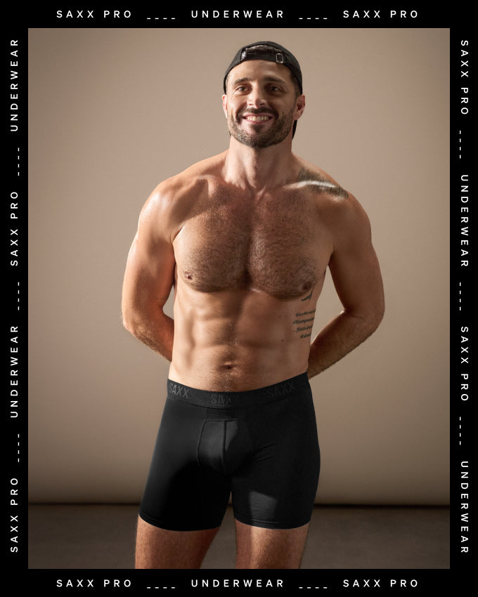 A shirtless man wearing black SAXX PRO underwear stands against a warm beige studio background, with SAXX PRO UNDERWEAR text repeated along the black border around the image.