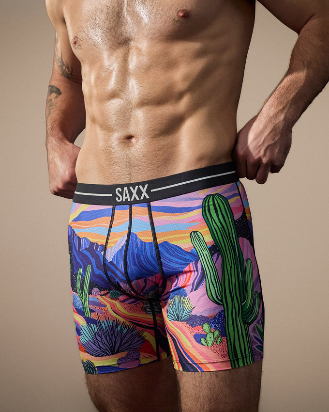 A close-up of a man's lower torso wearing SAXX boxer briefs with a colorful desert landscape print featuring cacti, mountains, and a pink, purple, blue, and orange sky.